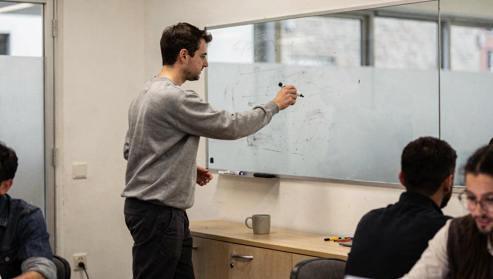 An engineer holding a marker points to a glass whiteboard while turning to speak with a seated colleague.