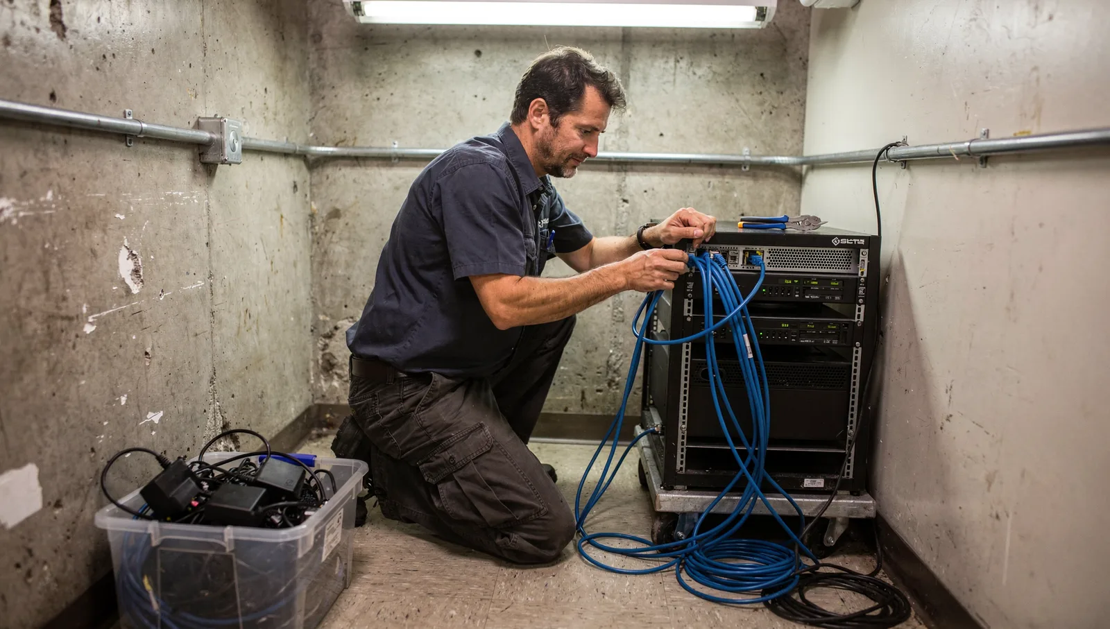 A systems engineer crouches by a wheeled server rack in a utility closet, untangling a bundle of blue Ethernet cables.