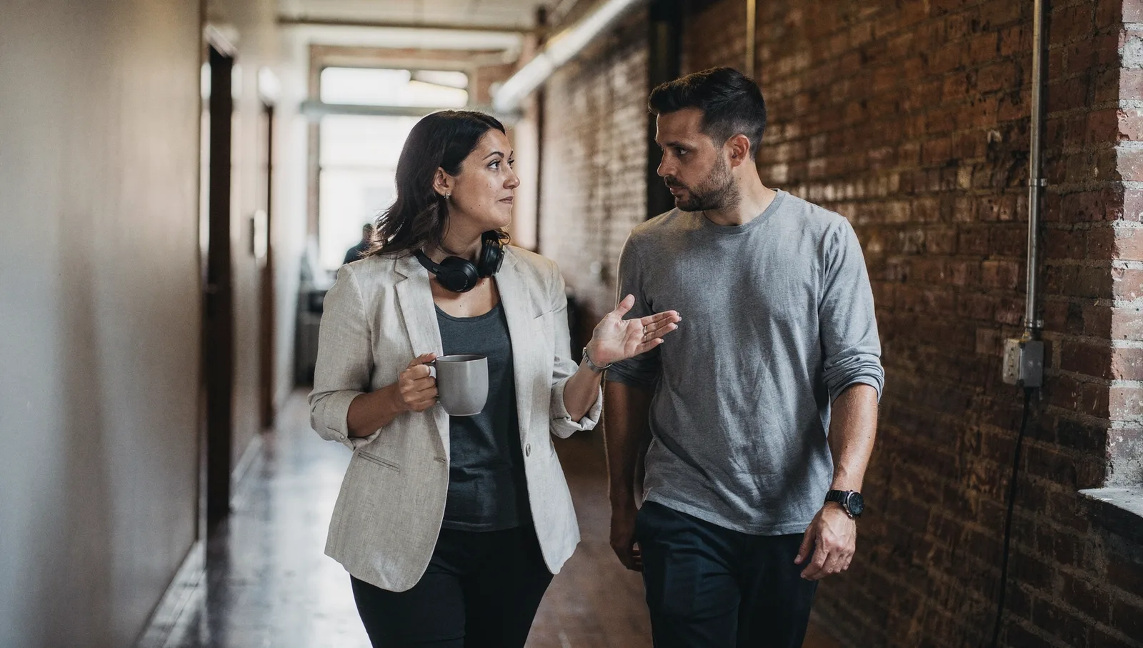 Two coworkers converse while walking down a bright office hallway with exposed brick walls.