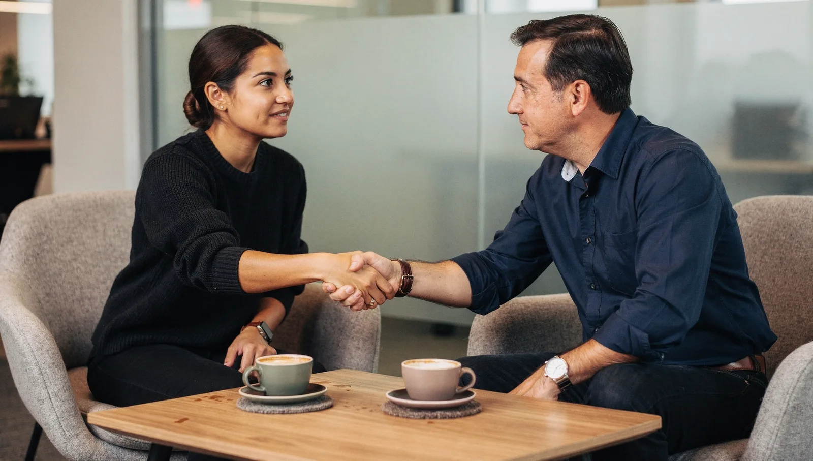 A woman and a man shaking hands over a low wooden coffee table in a modern lounge, with two espresso cups nearby.