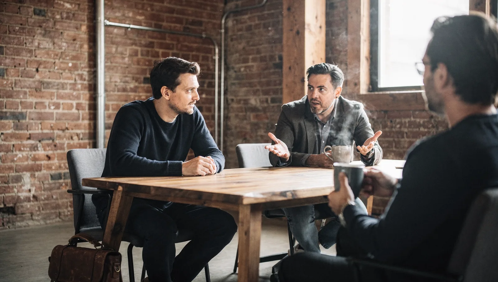 Two professionals seated at an oak table in a brick conference room, talking over mugs of coffee.
