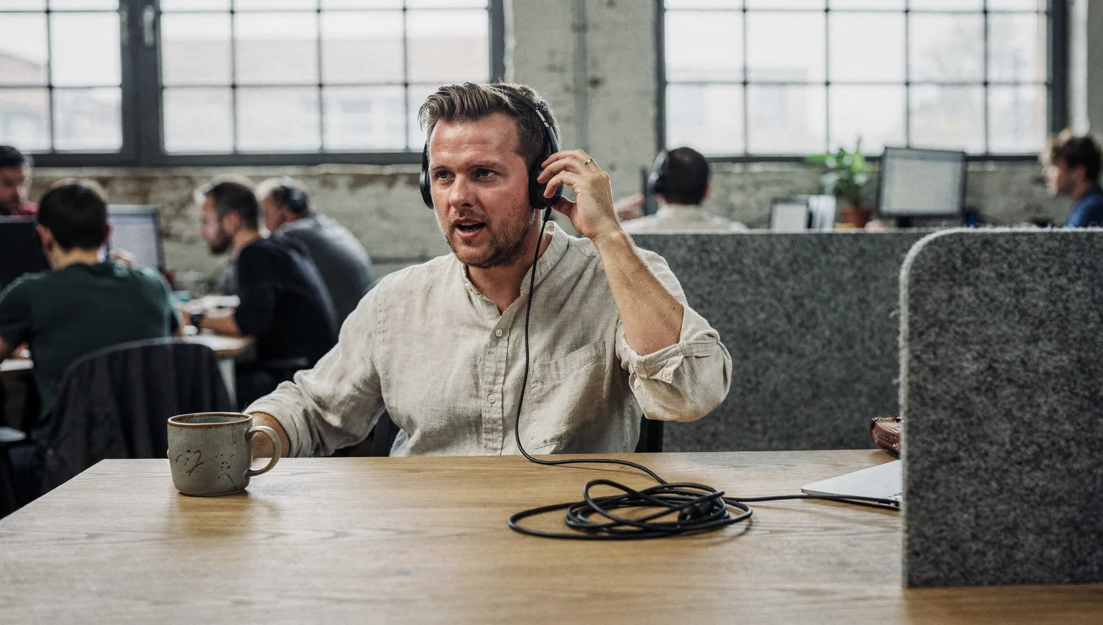 A person wearing a headset speaks at a wooden standing desk in an open-plan software agency office.